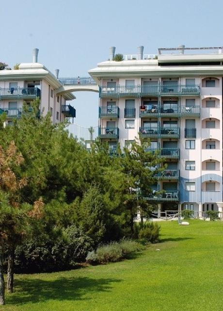 Dva moderní, několika podlažní obytné domy spojené lávkou s trávníkem a stromy v popředí.